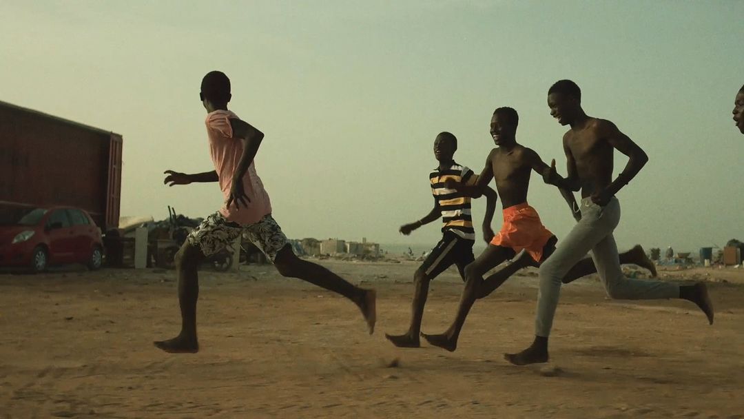 Group of African boys running barefoot on sandy terrain near the beach and buildings under a bright, overcast sky.