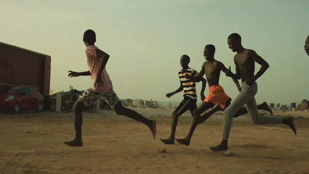 Group of African boys running barefoot on sandy terrain near the beach and buildings under a bright, overcast sky.