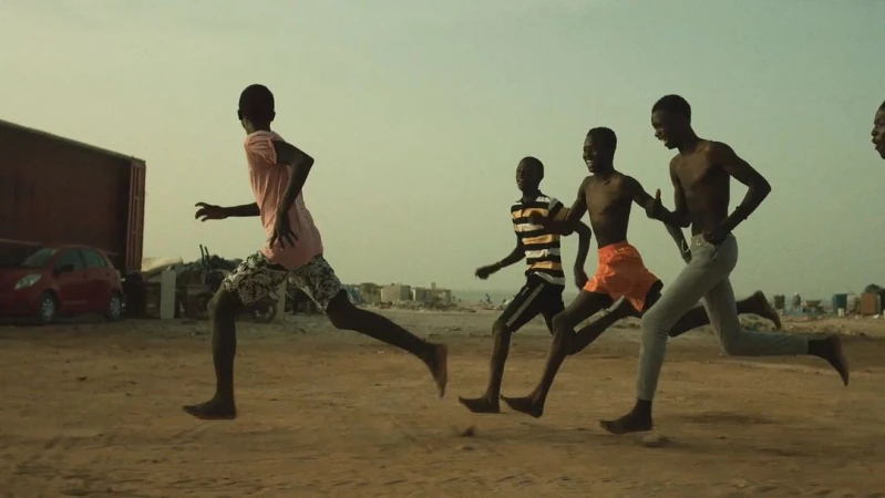 Group of African boys running barefoot on sandy terrain near the beach and buildings under a bright, overcast sky.