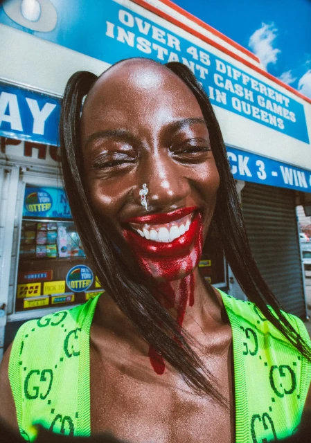 Woman with dramatic red makeup, closed eyes, and a bright green Gucci top stands in front of a lottery sign. Close-up portrait.