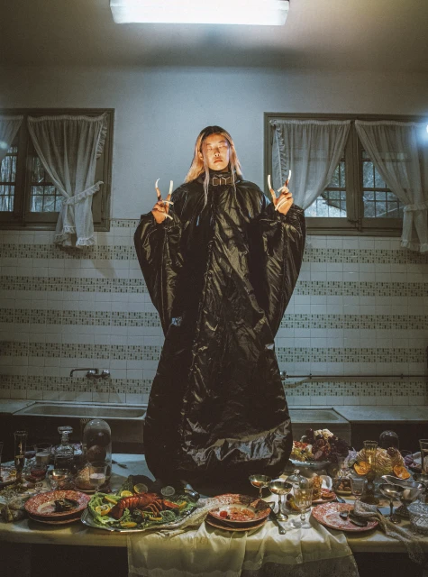 Portrait of a person in an oversized black coat standing at a table laden with food and drink in an indoor tiled environment.