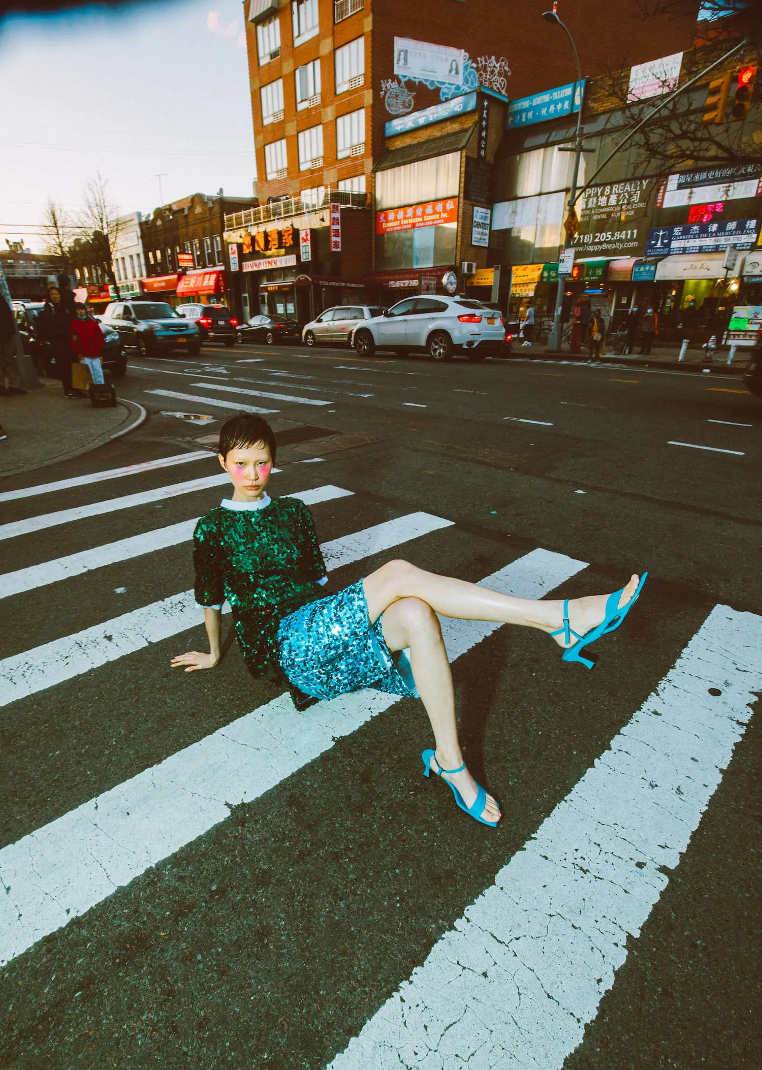 A female model poses on a zebra crossing in the city, wearing a green sequin top, blue skirt, and blue sandals on a sunny day.