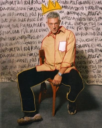 Portrait of Vincent Cassel seated on a wooden chair against a wall covered in handwritten script with a drawn yellow crown above him.