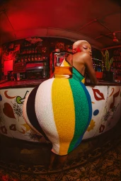 Woman in a striped multi-color dress leaning against a bar counter with a glass of amber liquid, shot with a fisheye lens.