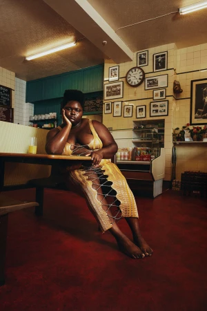 Plus-size black woman seated at a diner table wearing a yellow lace-up dress, photographed by Lou Escobar.