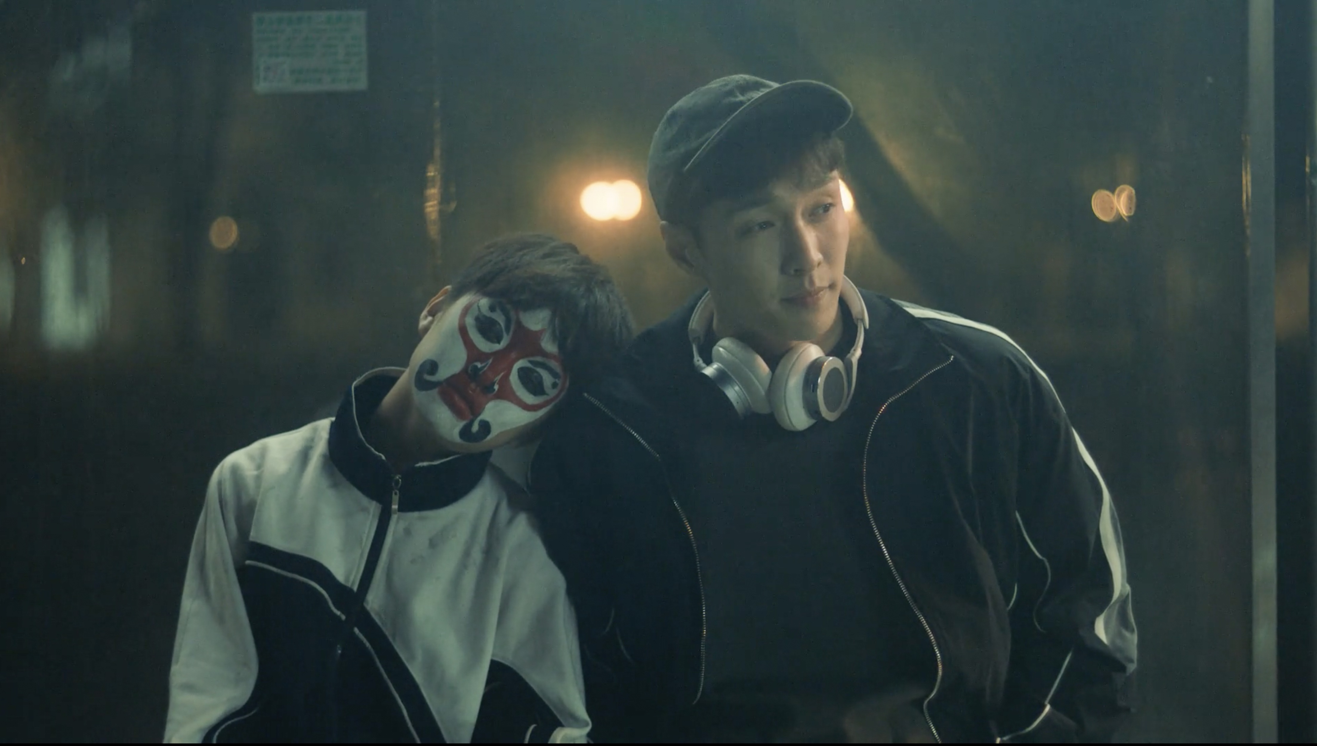 Two Asian young adult males in casual clothing, one with face paint, leaning against a dark background with bokeh lights.