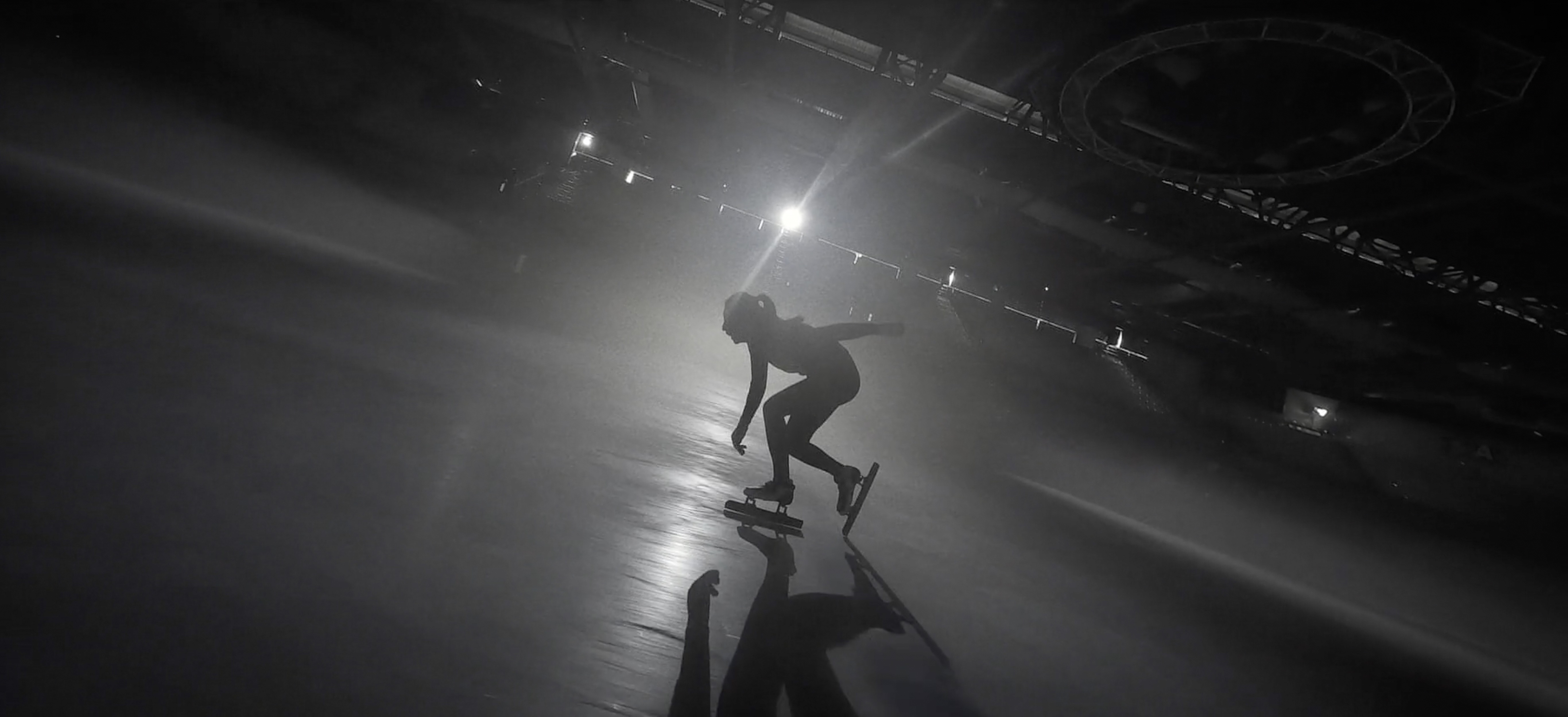 Monochrome shot of a speed skater in action on an indoor rink, captured from a high angle with dramatic lighting and reflection.