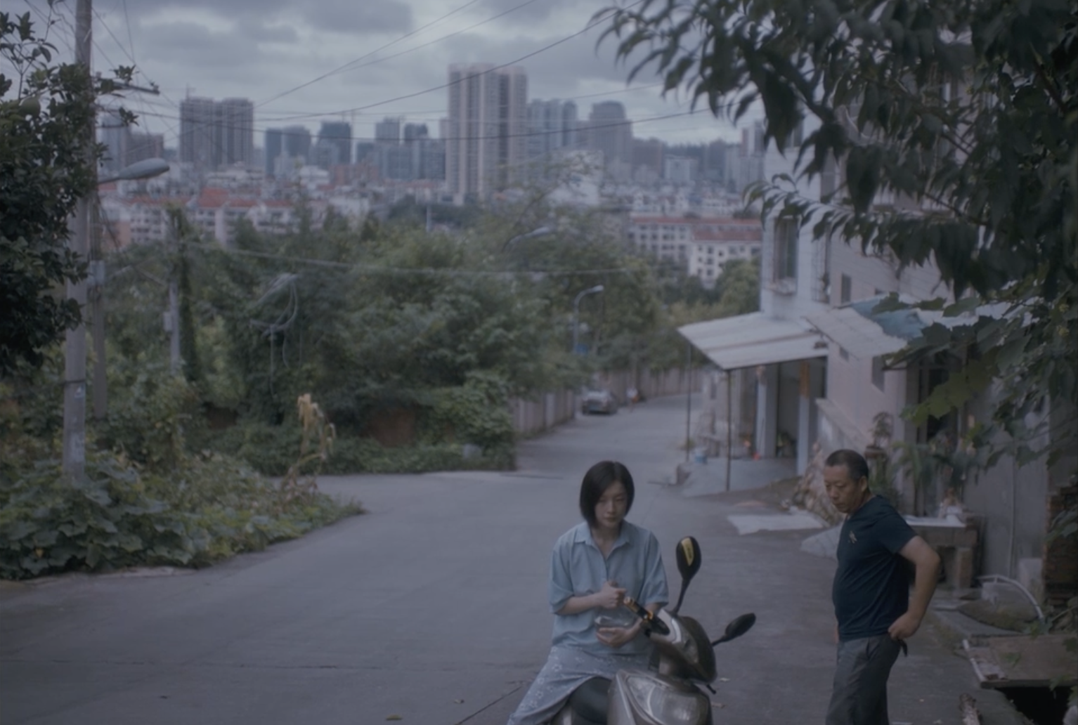 Woman on moped eating food on city street with an older man standing near. Tall buildings and green trees in the distant background.