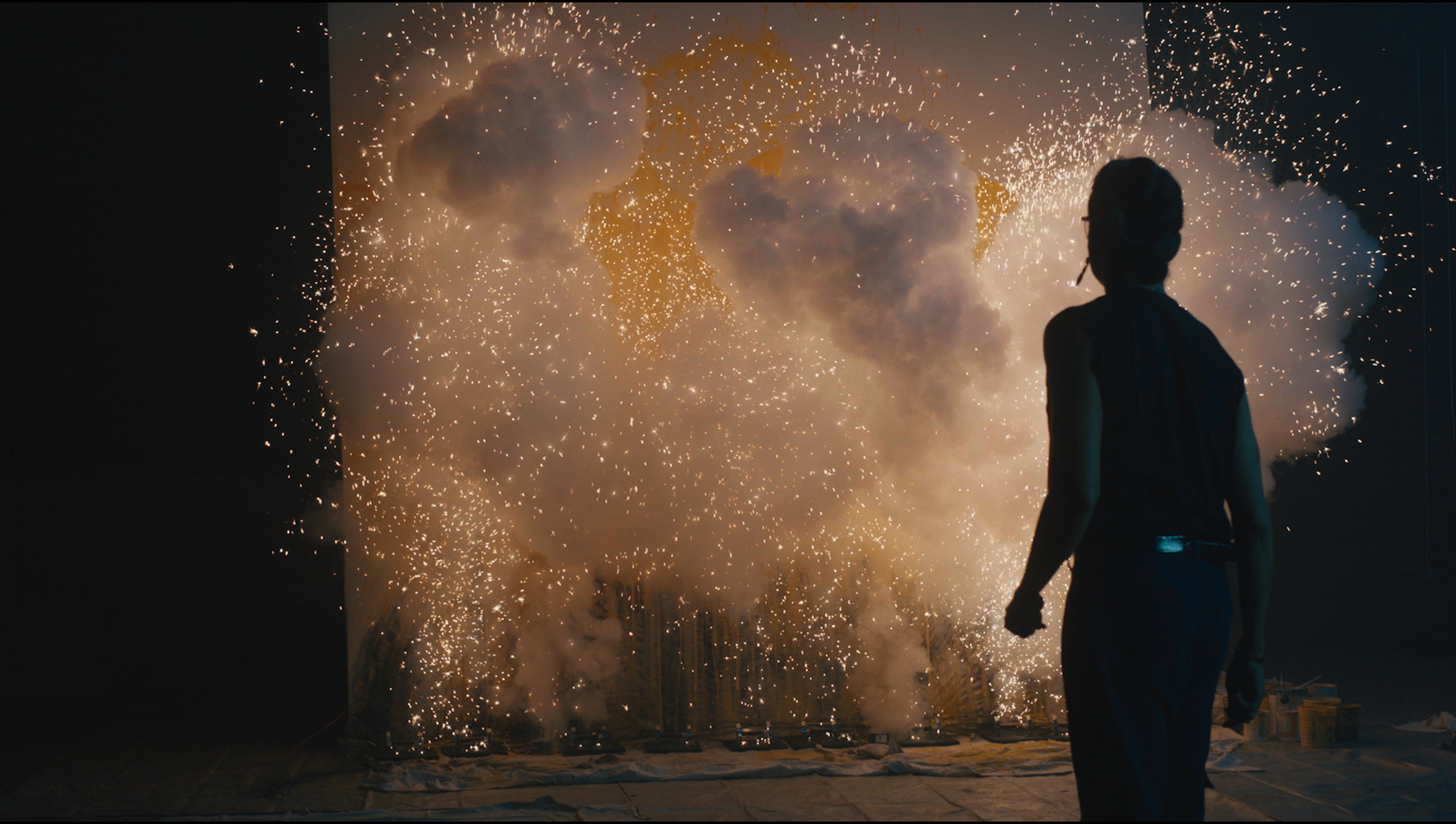 A person stands in front of a wall of sparks and smoke in an interior space, illuminated by golden light from the special effect.