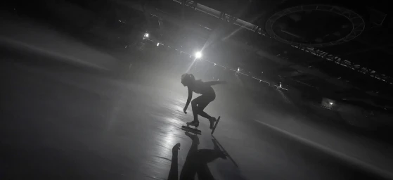 A silhouetted figure ice skating on an indoor rink under dramatic overhead lighting, captured at a low angle.