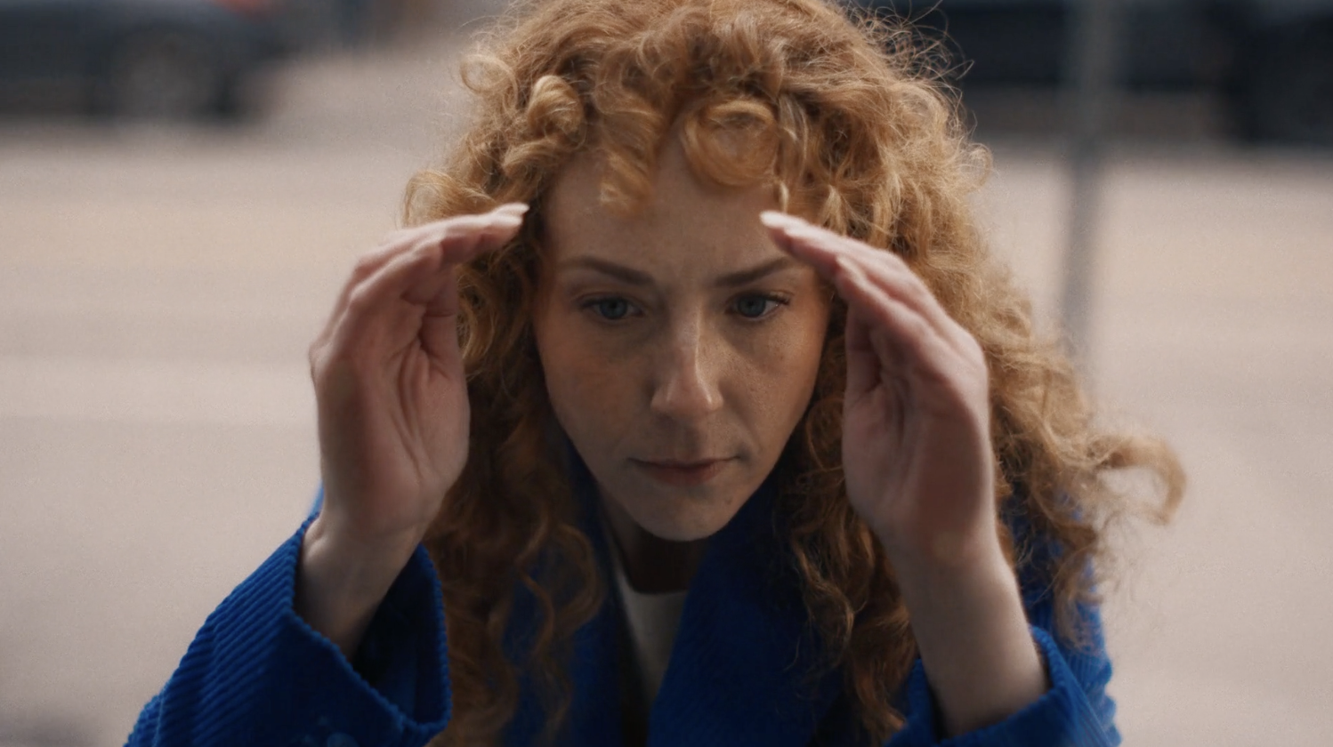 Close-up of a redhead woman in a blue coat touching her forehead with her fingertips, looking downwards in an outdoor urban setting.