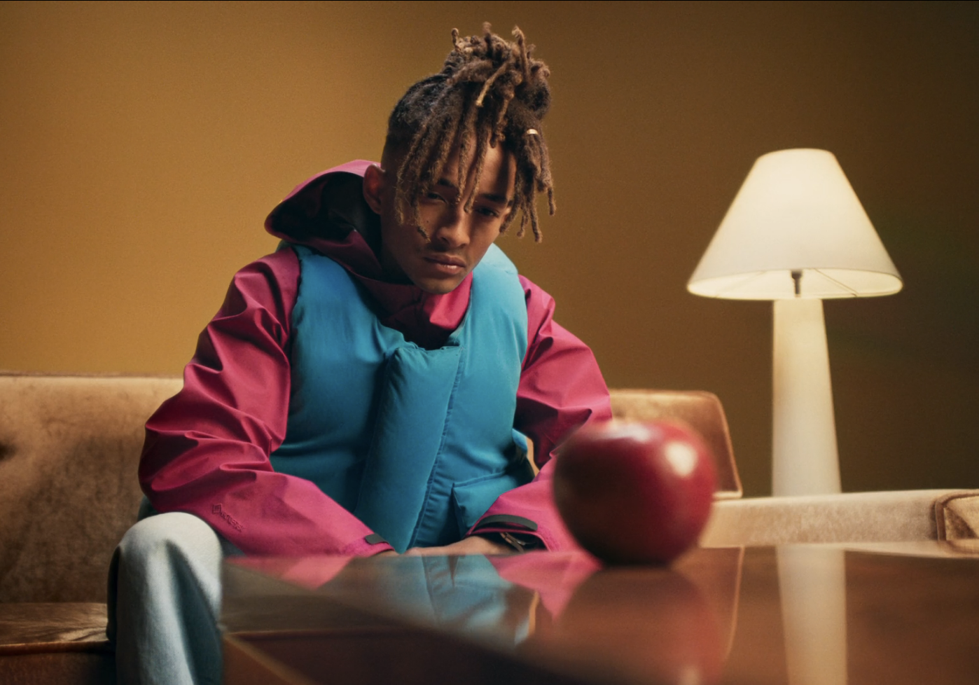 Young adult with dreadlocks sits near a red apple indoors, wearing a blue vest and pink jacket in front of a tan background and table lamp.