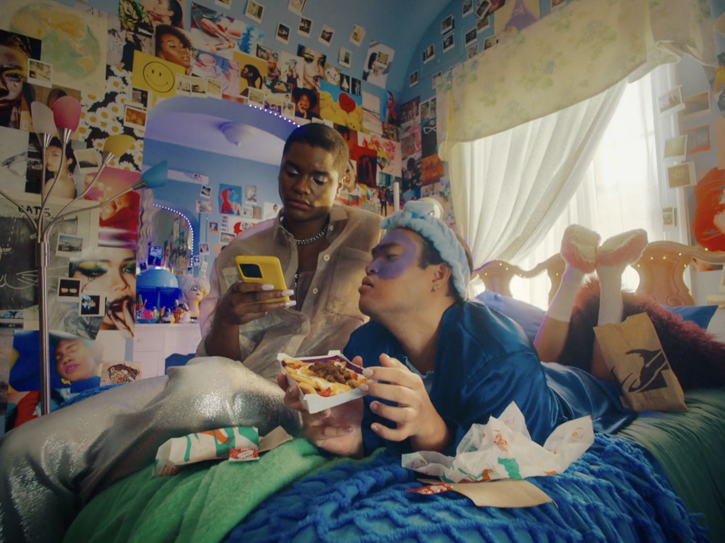 Two individuals lounge on a bed covered with snacks. One uses a phone, while the other eats from a food container in a casual indoor setting.