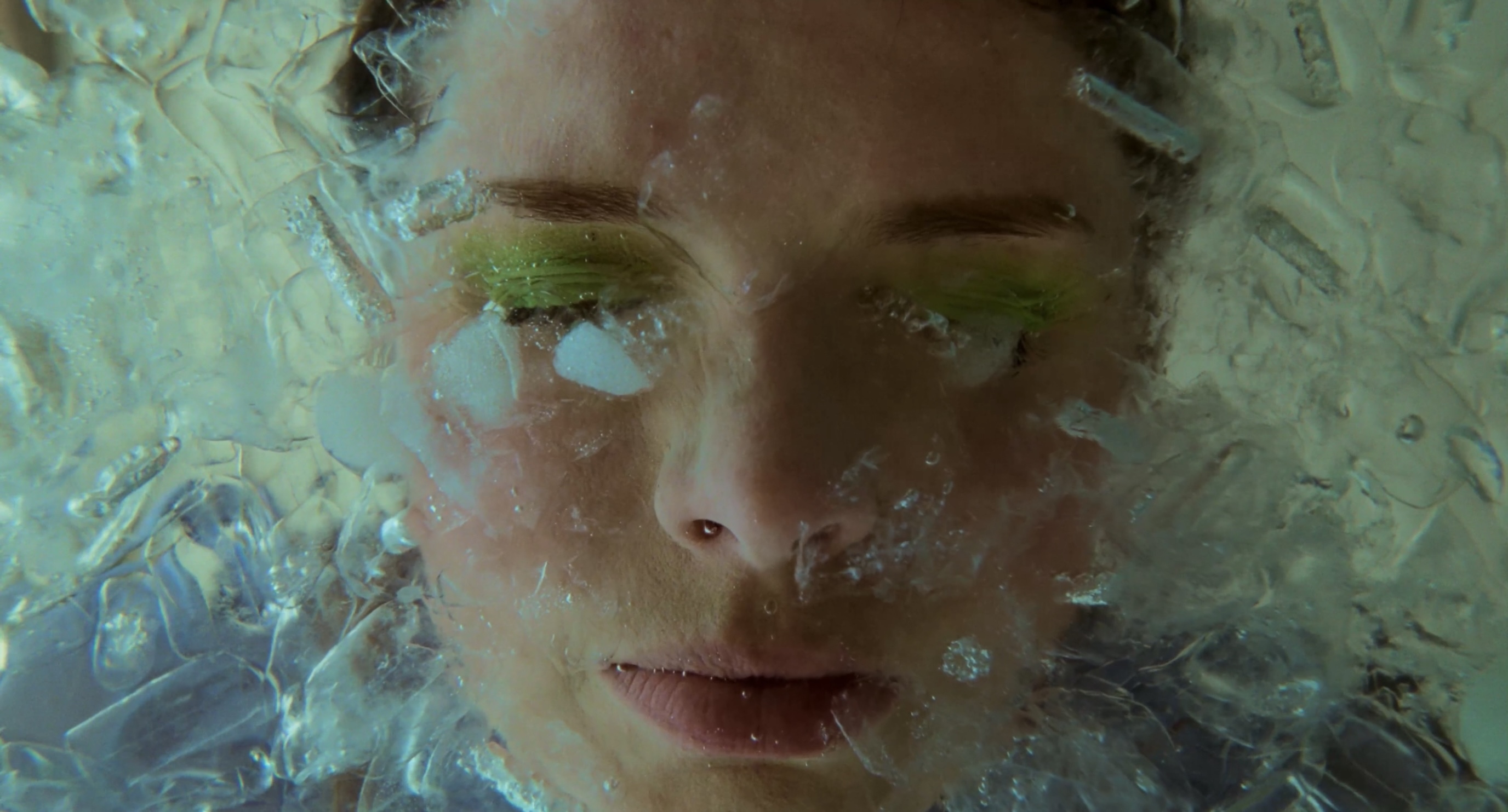 Close-up of a woman's face partially obscured by ice, featuring green eyeshadow. The image captures the interplay of beauty and cold.