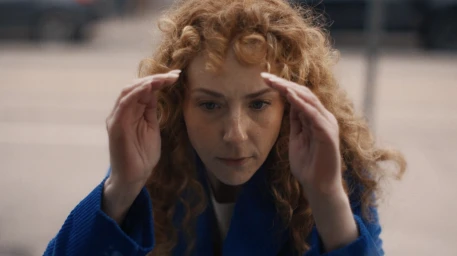 Close-up of a woman with curly red hair wearing a blue coat, holding her hands up near her forehead outdoors.