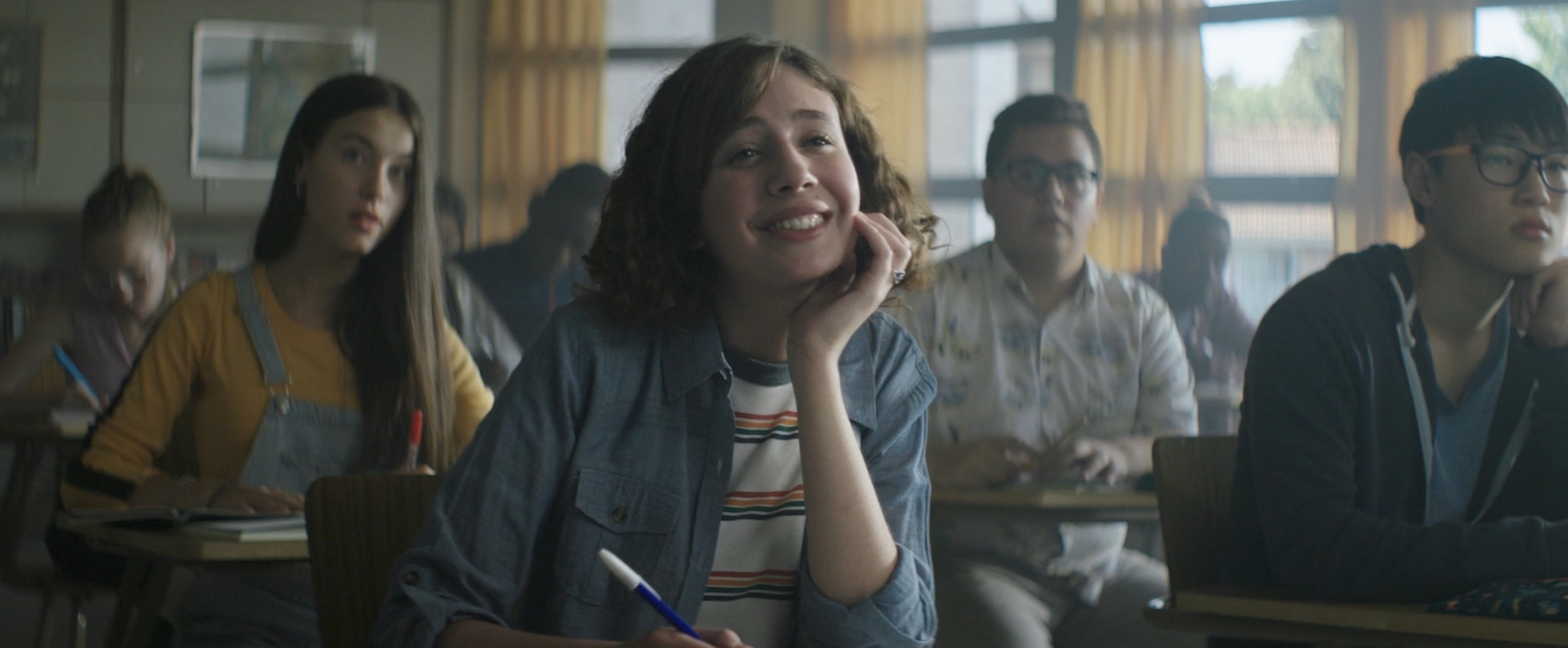High school classroom setting with smiling student in the foreground, classmates and desks filling the background, natural light.