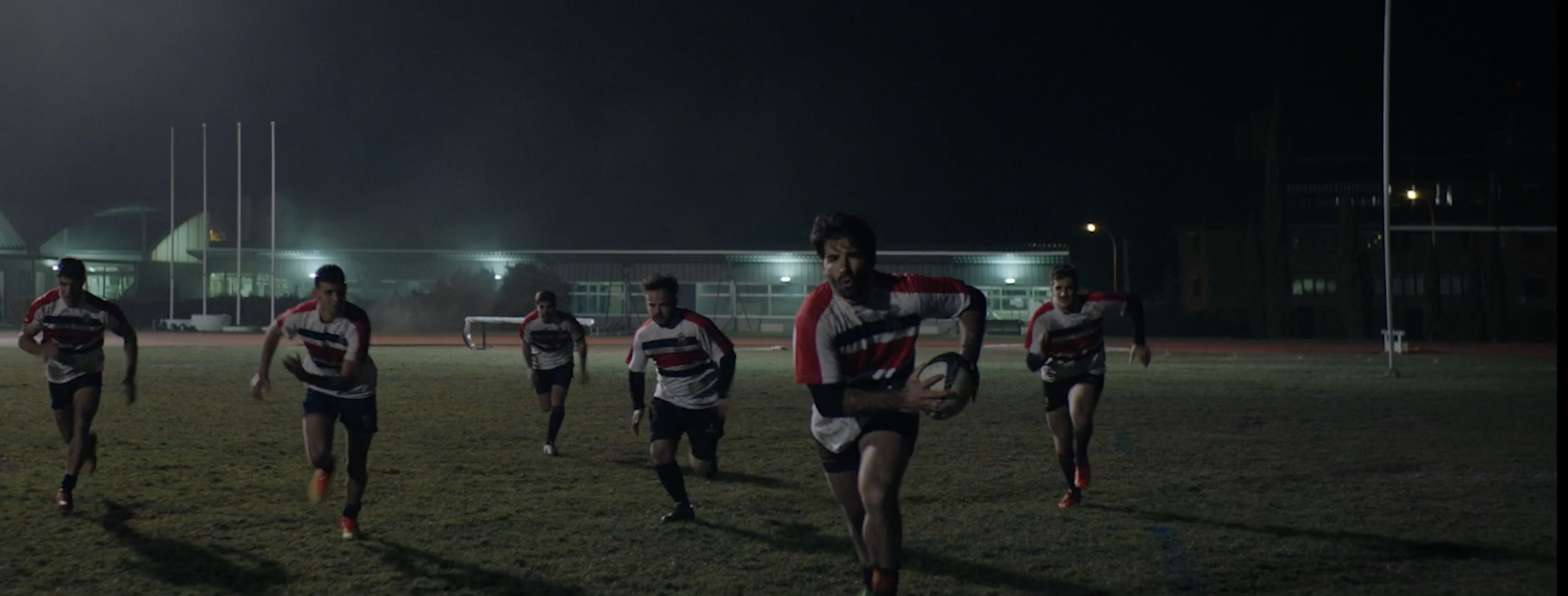 Rugby players running on a field at night, wearing red, white, and black jerseys, carrying a rugby ball, and playing under the lights.