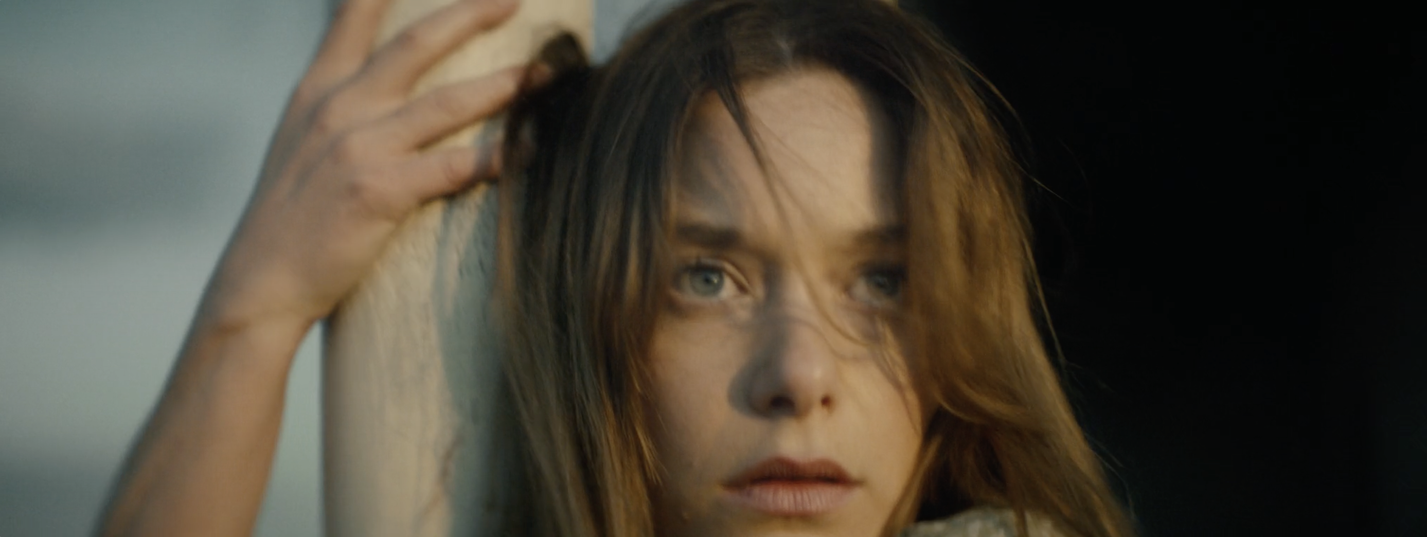 Close-up portrait of a young woman with long brown hair resting her hand against a pillar outdoors.