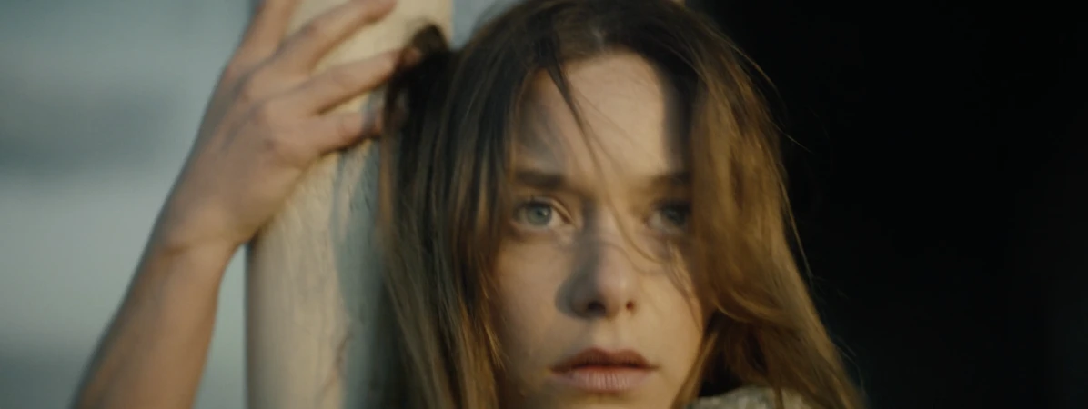 Close-up portrait of a young woman with long brown hair resting her hand against a pillar outdoors.