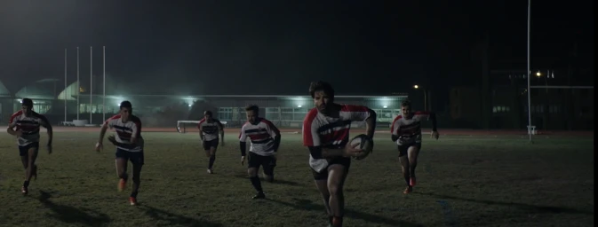 Rugby players running across a floodlit sports field at night carrying a ball, filmed by Miguel Campana in Madrid.