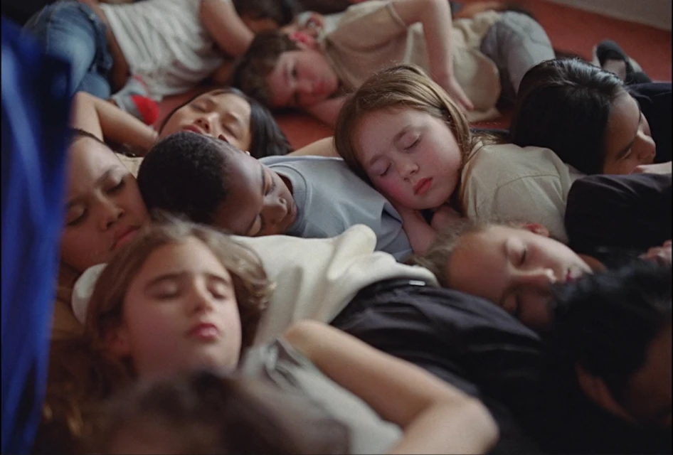 Group of children of various ethnicities lying close together, napping indoors on a red floor, illuminated by soft, natural light.