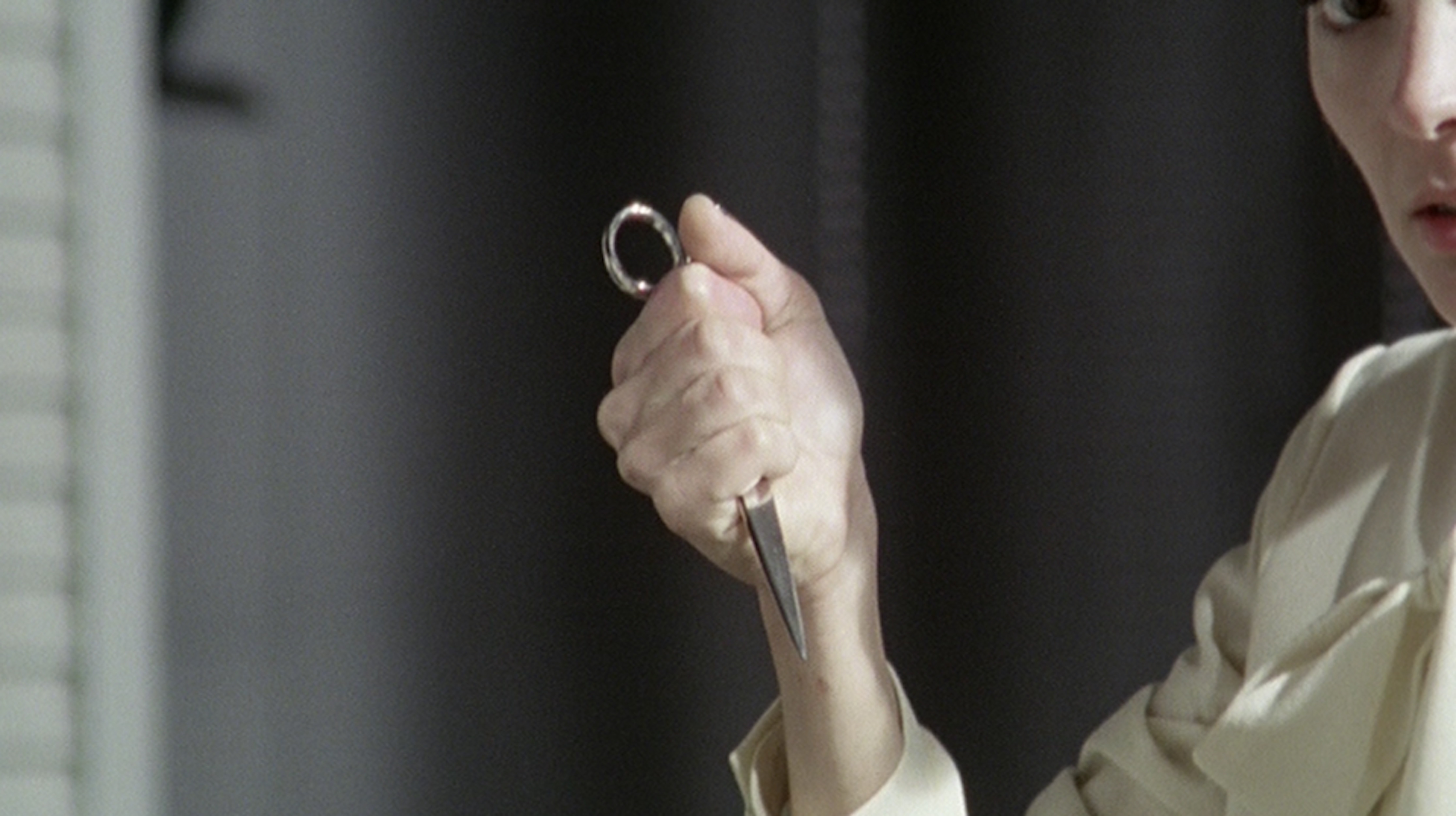 Close-up of an adult holding a small metal scissors, with a neutral facial expression, in an indoor environment against a gray background.