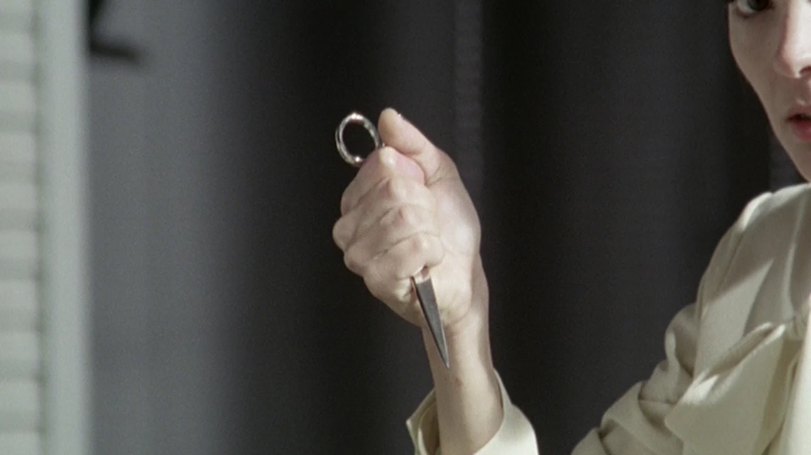 Close-up of an adult holding a small metal scissors, with a neutral facial expression, in an indoor environment against a gray background.