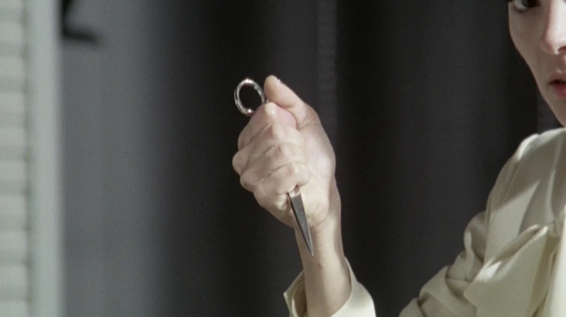 Close-up of an adult holding a small metal scissors, with a neutral facial expression, in an indoor environment against a gray background.