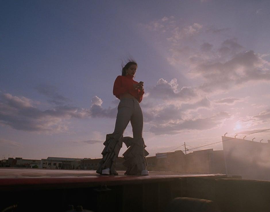 A young adult woman stands on a rooftop in flared pants, using her phone with a cloudy sky and buildings in the background.