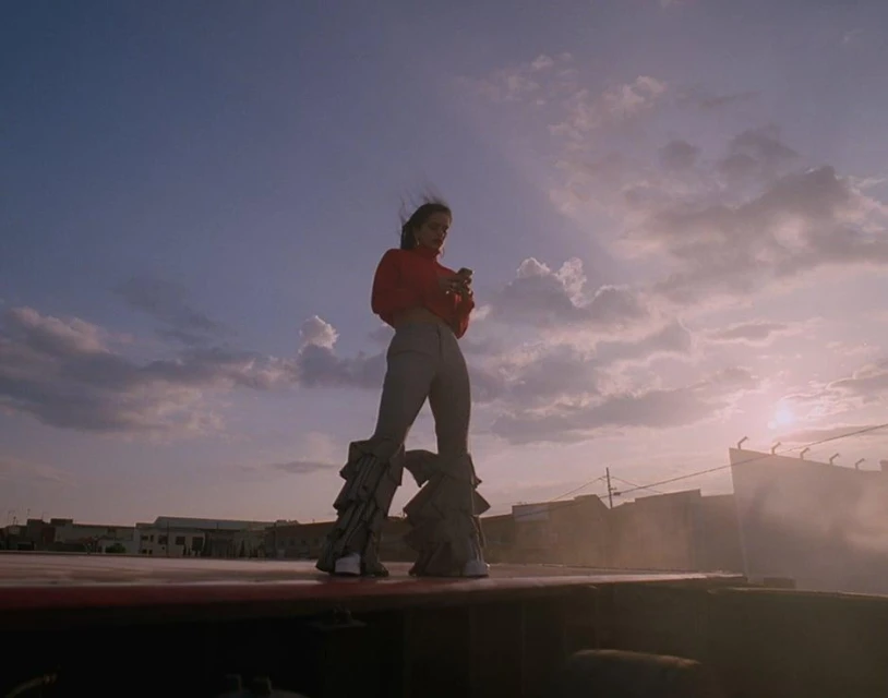 A young adult woman stands on a rooftop in flared pants, using her phone with a cloudy sky and buildings in the background.