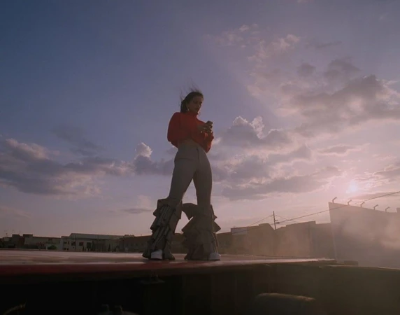 A young adult woman stands on a rooftop in flared pants, using her phone with a cloudy sky and buildings in the background.