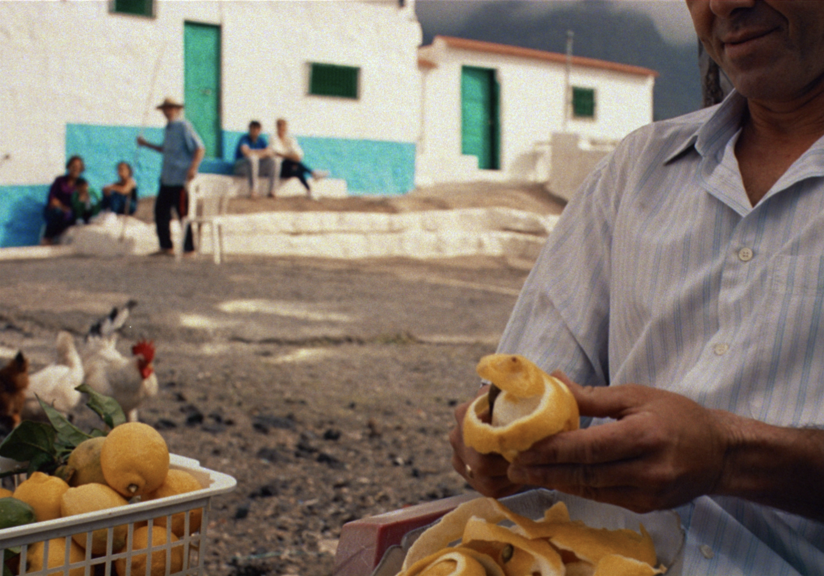 Man peels lemons at an outdoor market in a rural setting with a group of people and chickens visible in the background on a sunny day.