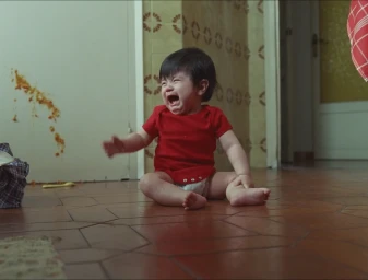 Crying baby wearing a red onesie seated on a terracotta tiled floor with food smeared on the wall behind