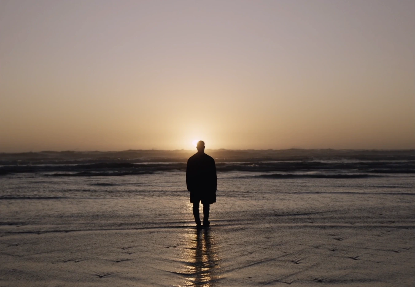 Silhouette of a man standing alone on a wet sandy beach facing a glowing sunset over the ocean.
