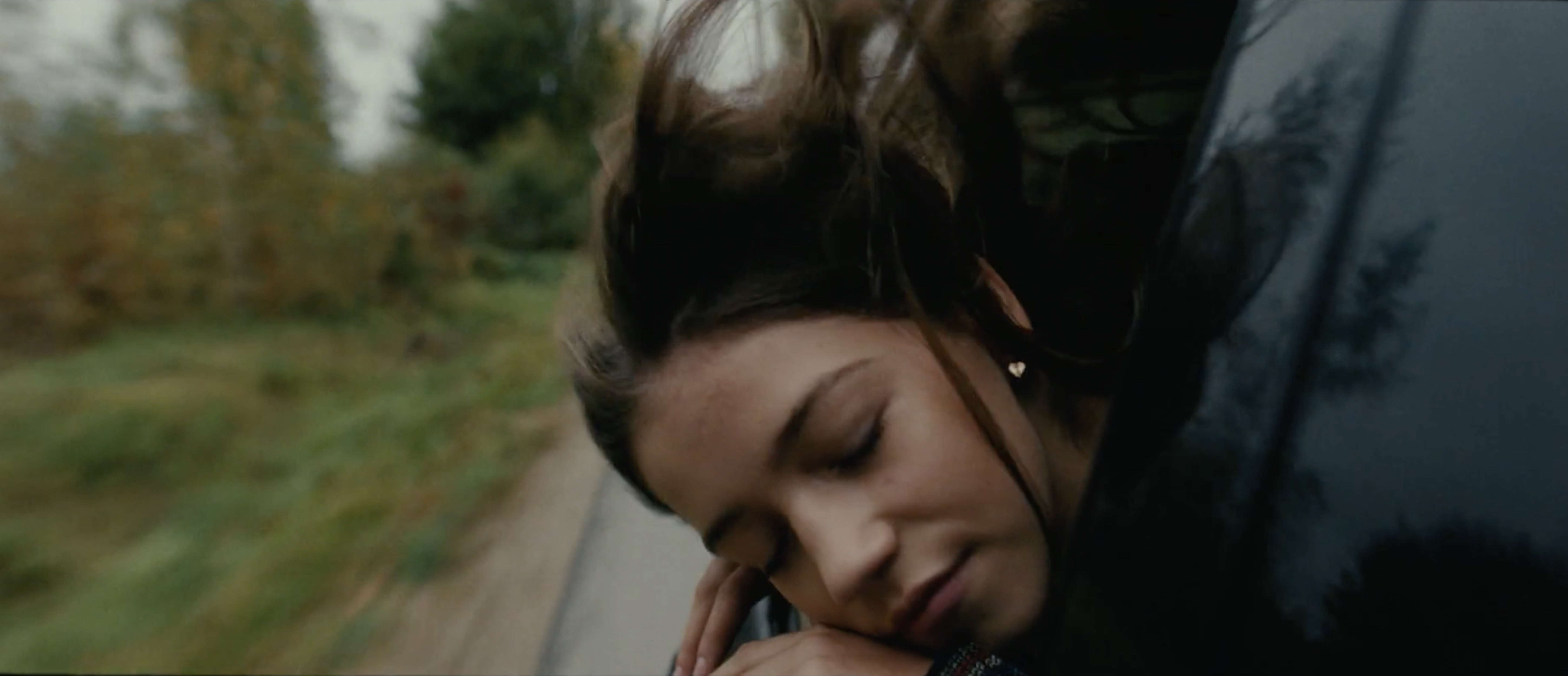 Young woman with closed eyes rests her head on her hand while sitting in a car with a landscape of trees in the background.