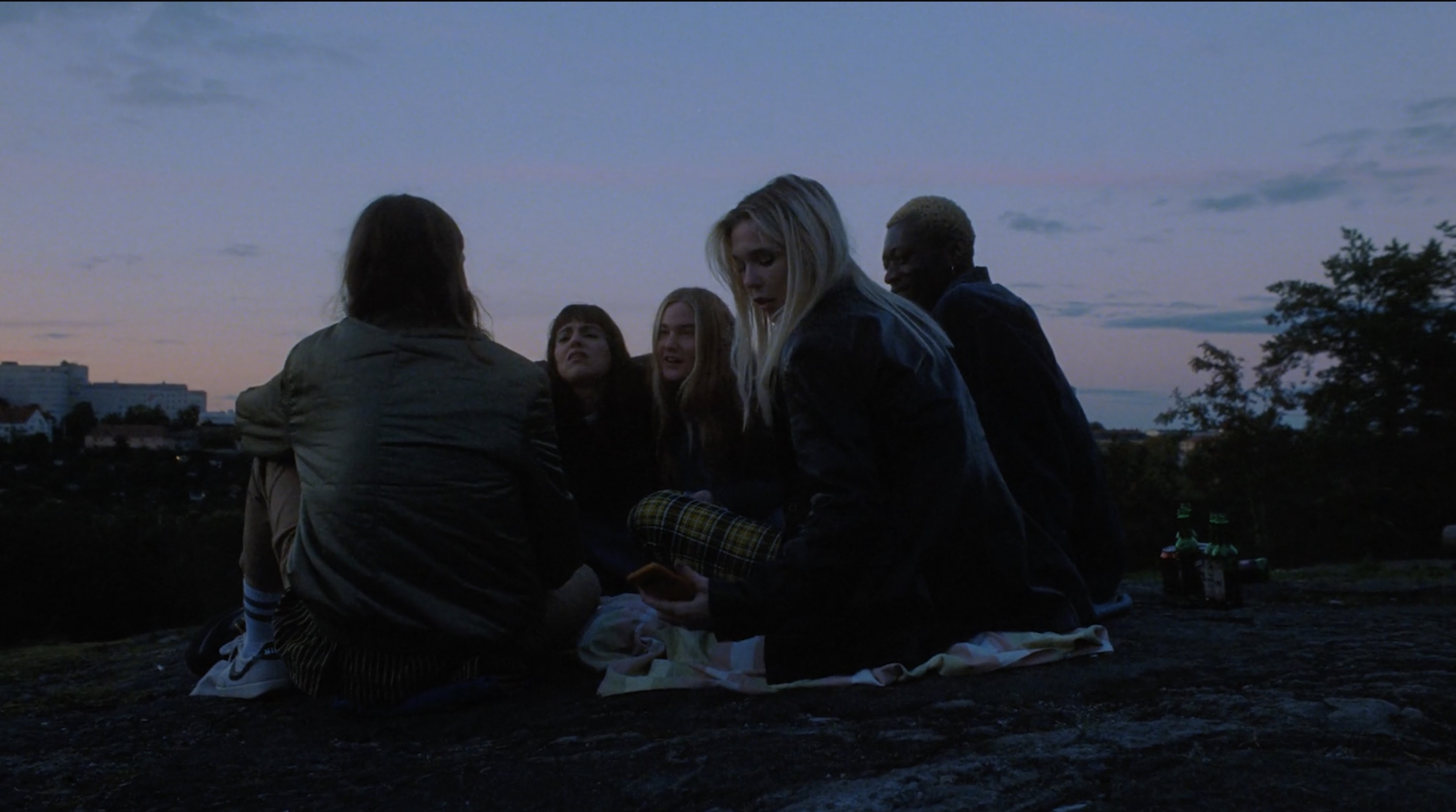 A group of five young adults sit on a rocky surface at dusk, with city buildings visible in the distance. The group is conversing.