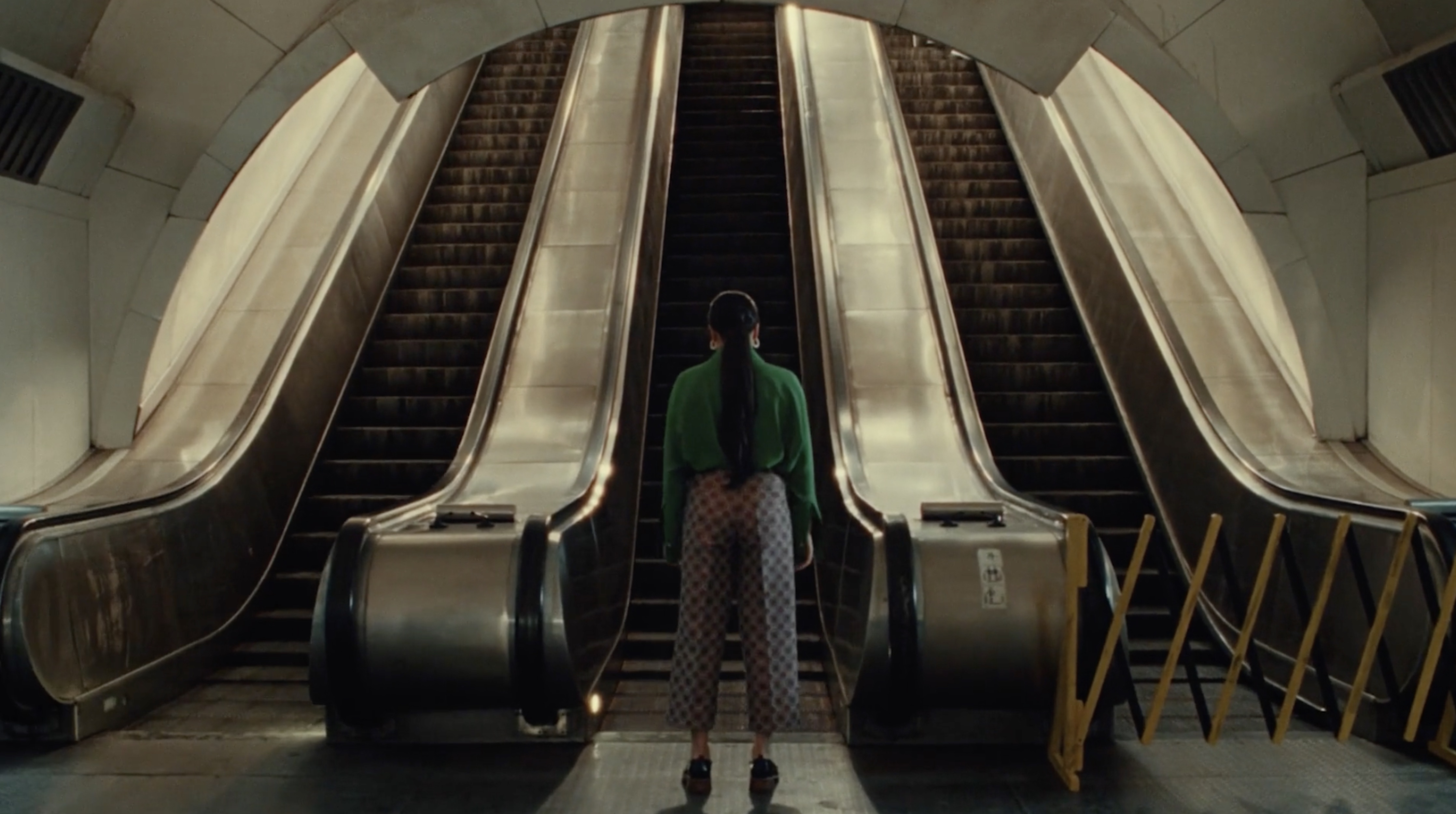 A young woman stands at the base of two escalators in an urban environment, her back to the camera, wearing a green shirt and patterned pants.