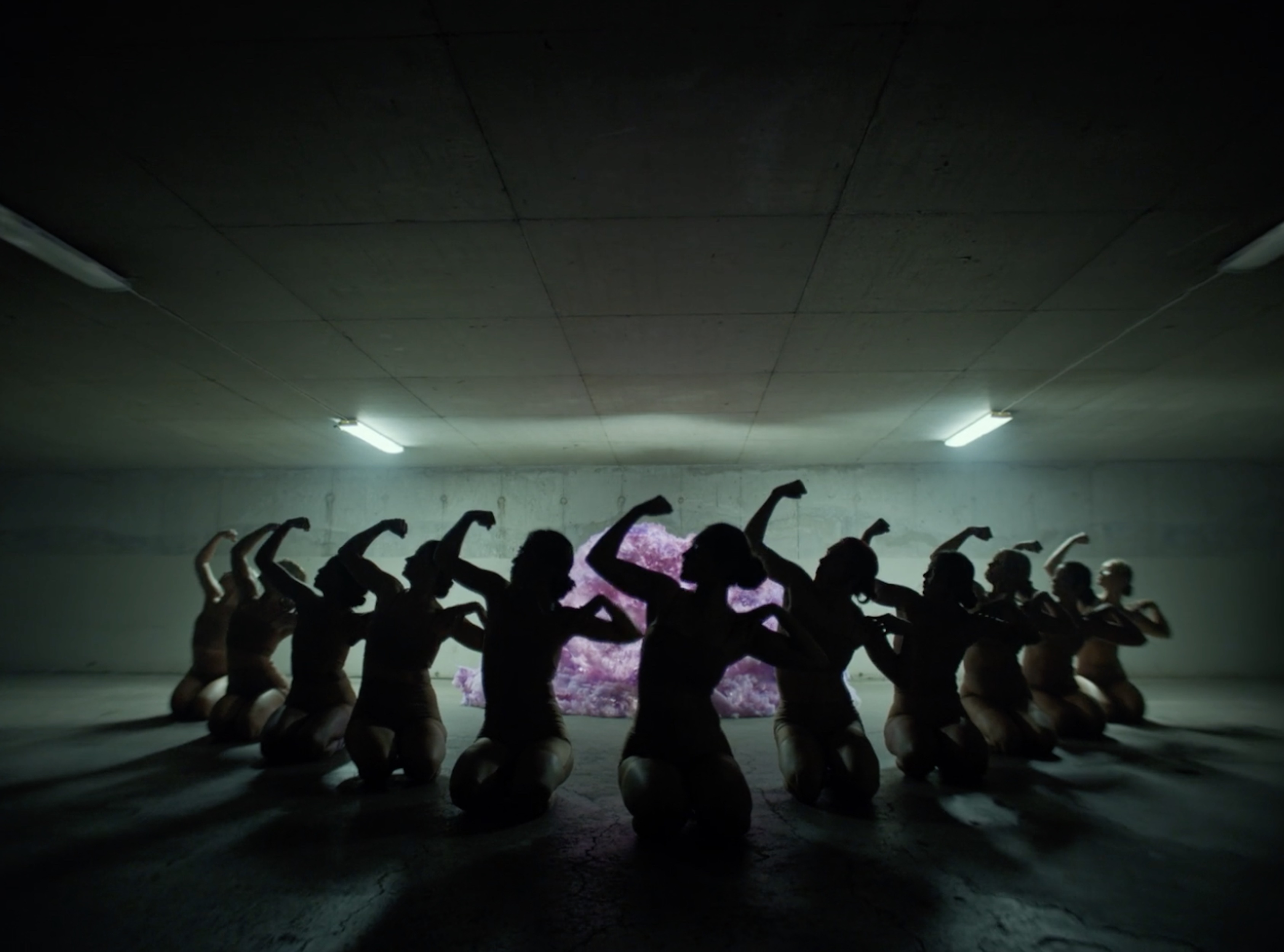 Group of individuals kneeling in dark tunnel beneath fluorescent lights, arms raised in unified gesture; possible performance art piece.