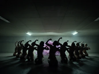 Silhouetted group of kneeling dancers in a dark industrial garage with a fluffy pink object in the background