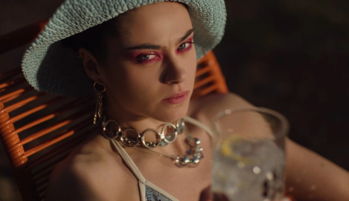 An adult female in a hat and jewelry sits in a chair holding a glass of water with a lemon slice, shot in medium view.
