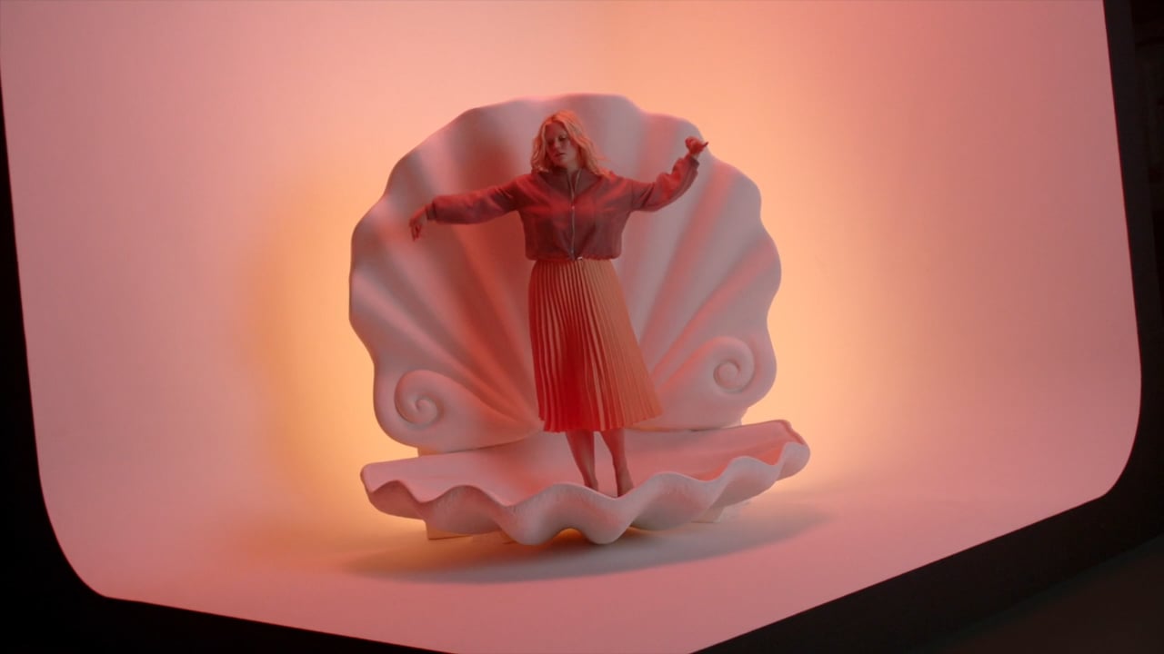 Adult female stands in a clam shell in a photo studio wearing a skirt and jacket in front of peach colored seamless backdrop.
