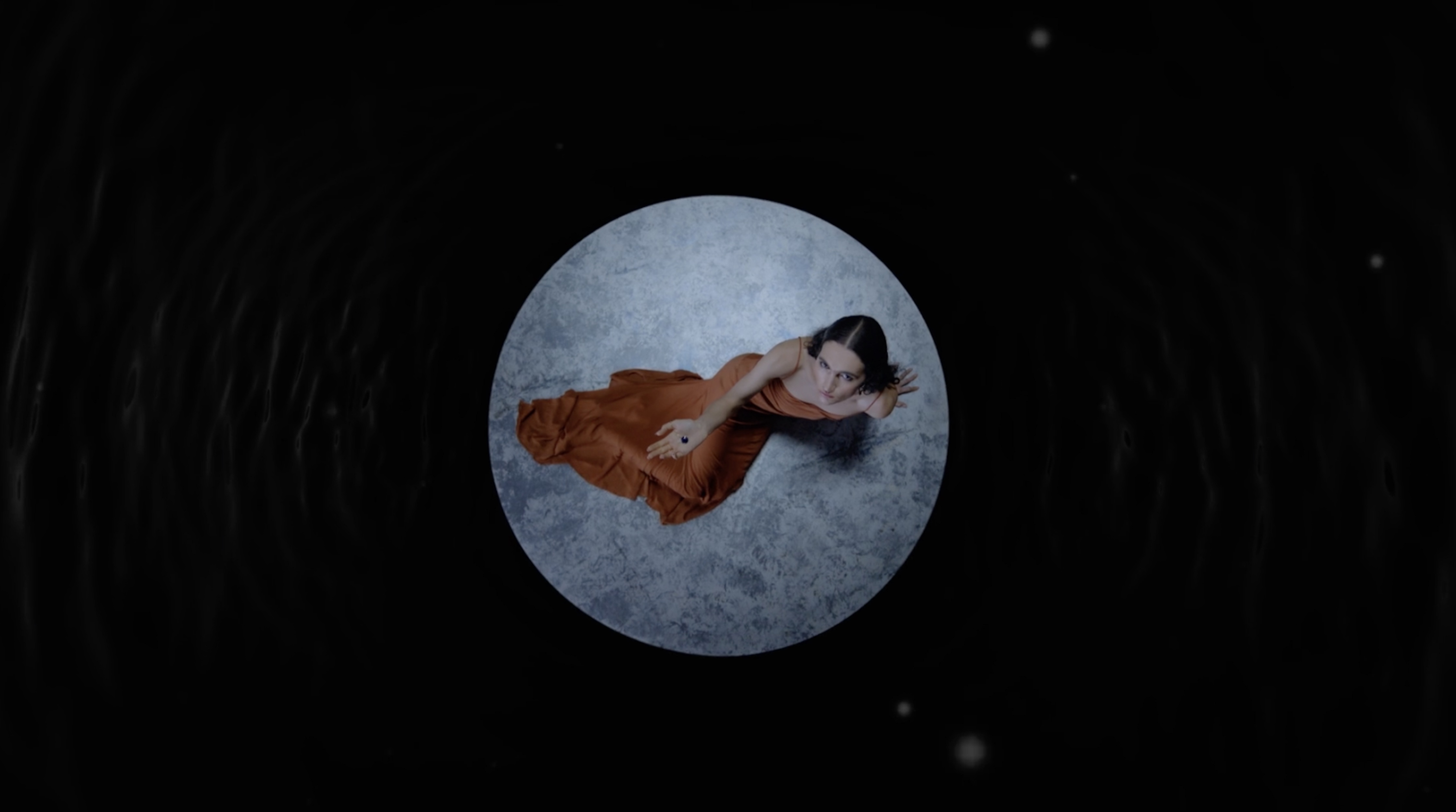Overhead shot of a woman in an orange dress lying on a gray circular surface surrounded by a dark, textured background with white specks.