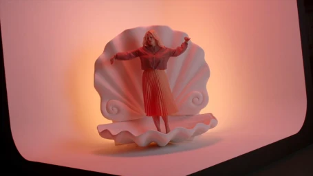 Woman in pink jacket and orange pleated skirt standing inside a giant sculptural white seashell prop on a curved studio backdrop