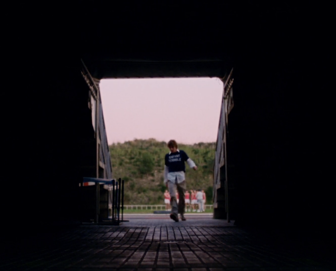 A young man walks through a dark tunnel leading to an outdoor track with other people, trees, and a pink sky visible in the distance.