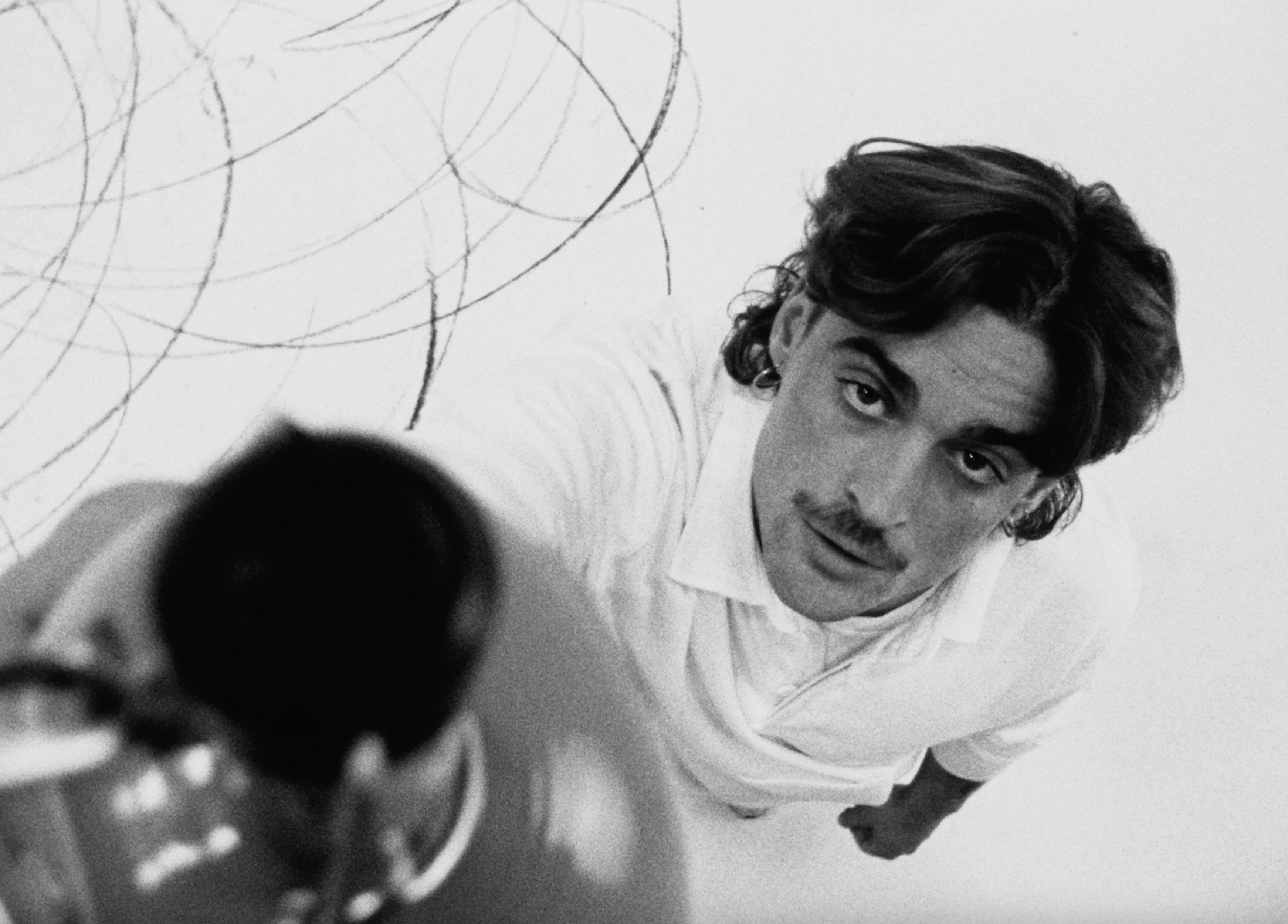 Low-angle portrait of an adult male with a mustache in a white collared shirt, looking up, with abstract pencil lines in the background.