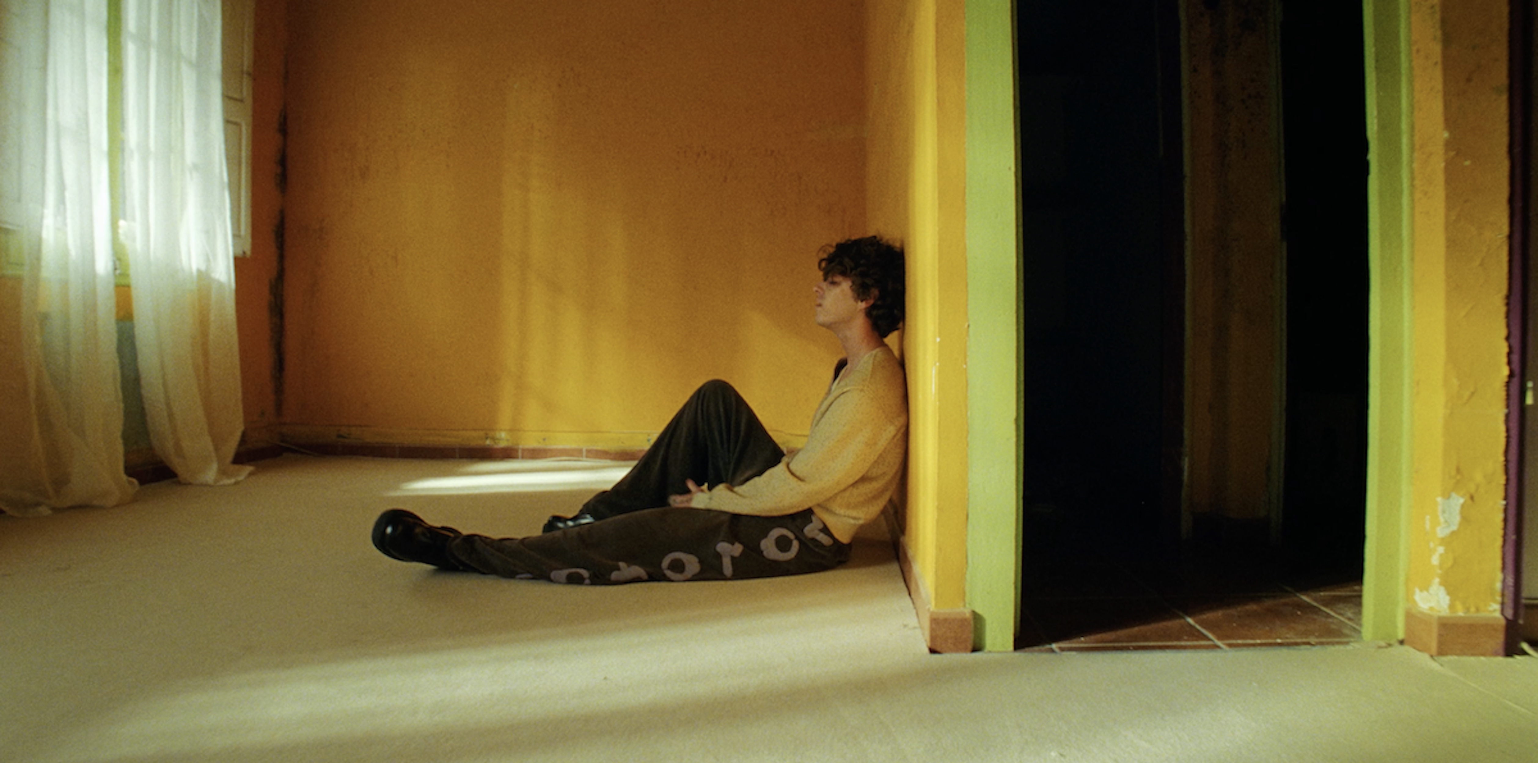 Young man sits in corner of yellow room lit by window light wearing casual sweater and pants with writing near dark doorway.