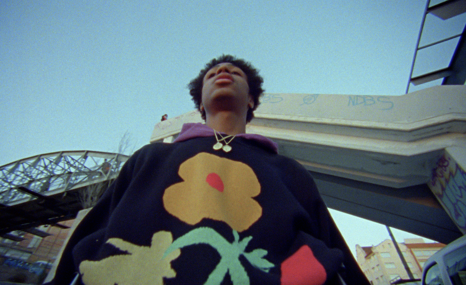 Low-angle shot of a young adult with an afro wearing a floral sweater standing in an urban setting, looking up at the sky and architecture.