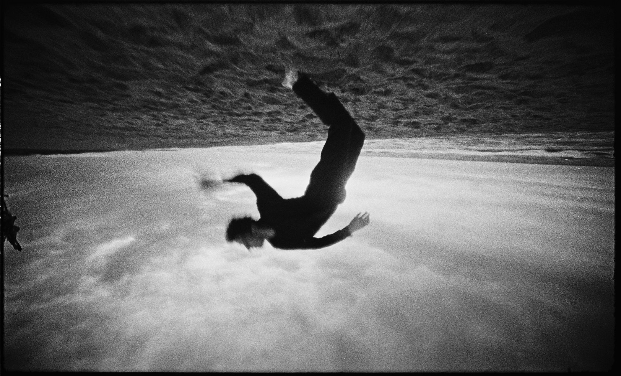 Monochrome image of an adult man in mid-air, falling backwards. He wears dark clothing, against a background of sky and a strip of ocean.