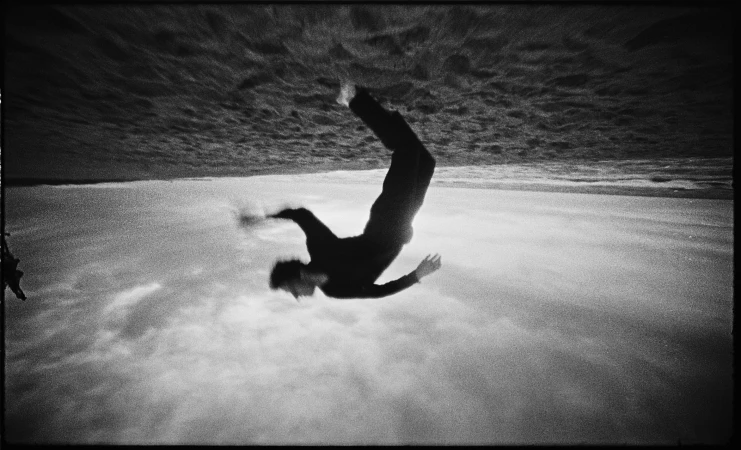 Monochrome image of an adult man in mid-air, falling backwards. He wears dark clothing, against a background of sky and a strip of ocean.