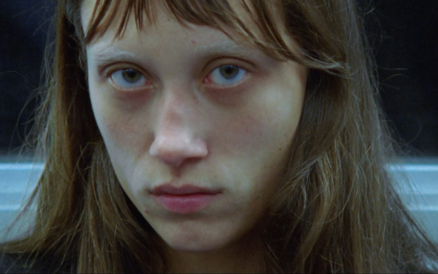Close-up portrait of a young adult female with light skin, brown hair, and a neutral expression against a blurred, dark background.